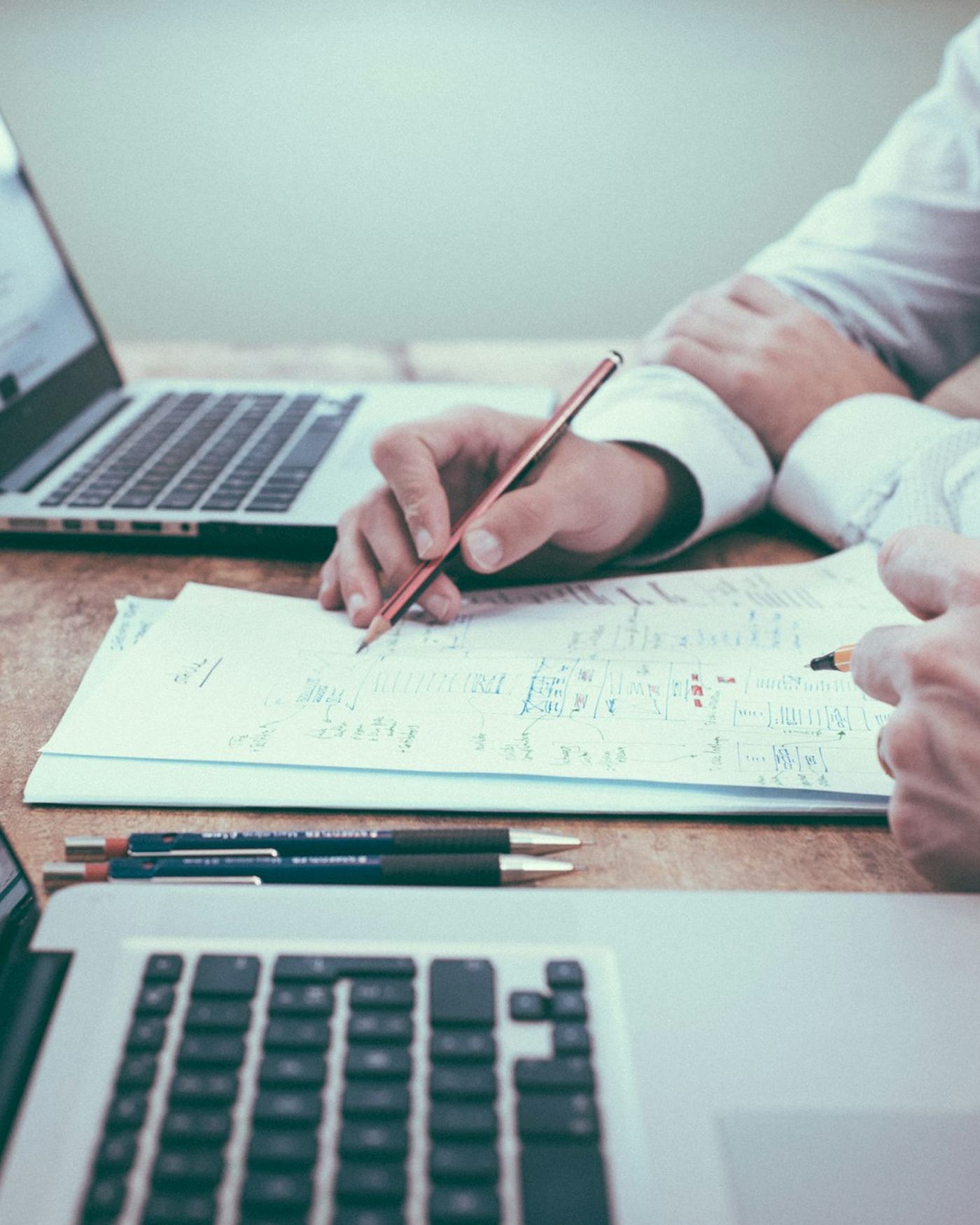 Manos revisan documentos y esquemas junto a computadores portátiles.